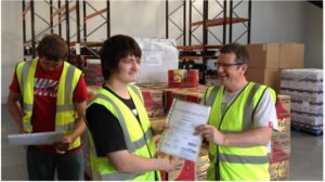 Three men in the FareShare warehouse; two wear high-visibility vests. One hands a certificate to another, while the third is behind them holding a paper. Photo credit: FareShare Thames Valley/SOFEA